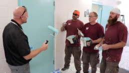 Inmates learning from construction program manager on how to properly use roller to paint walls